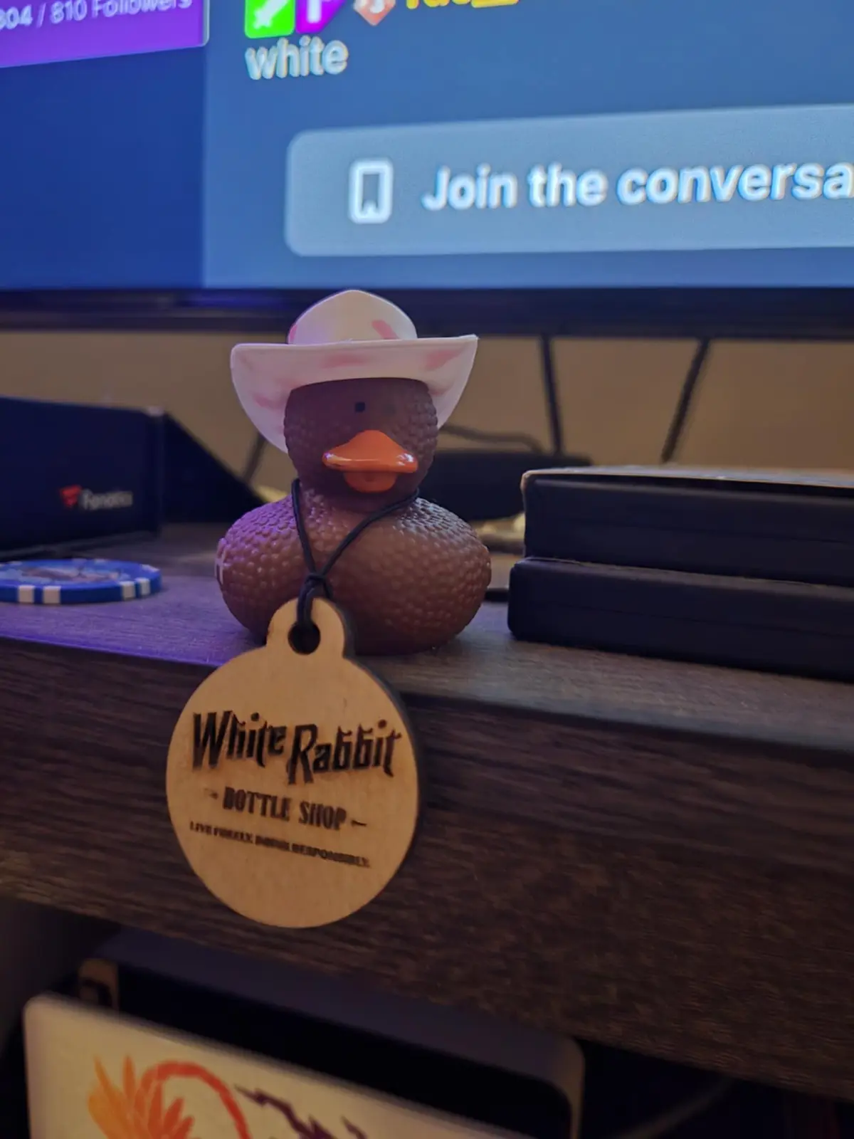 A brown rubber duck wearing a pink cowboy hat sits on a wooden bar counter, with a wooden tag reading 'White Rabbit - BOTTLE…
