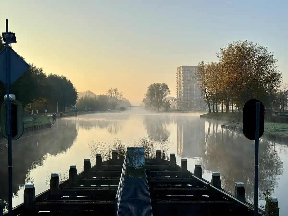 A serene, misty canal at sunrise with calm water reflecting the sky and distant buildings, flanked by trees and traffic…