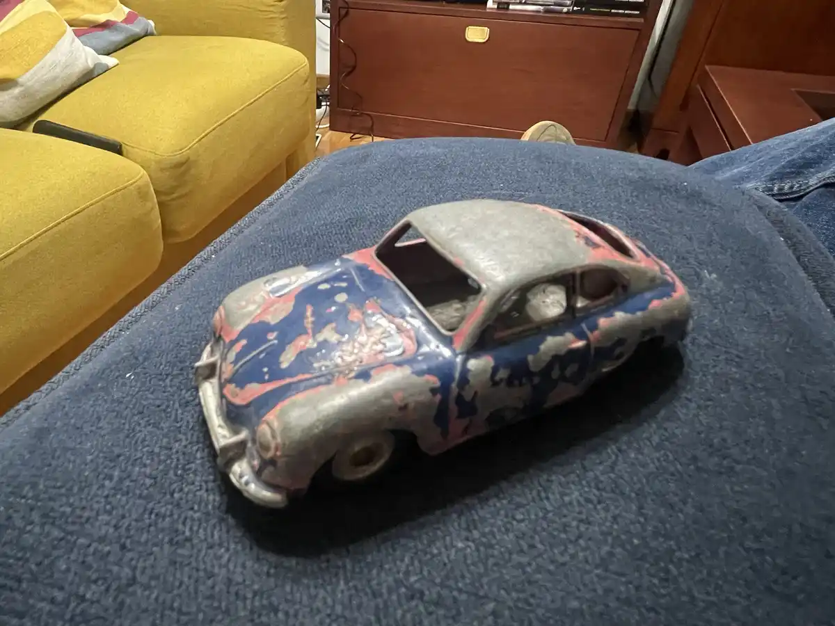 A heavily weathered and chipped toy car with peeling blue, red, and gray paint sits on a blue textured blanket. In the…