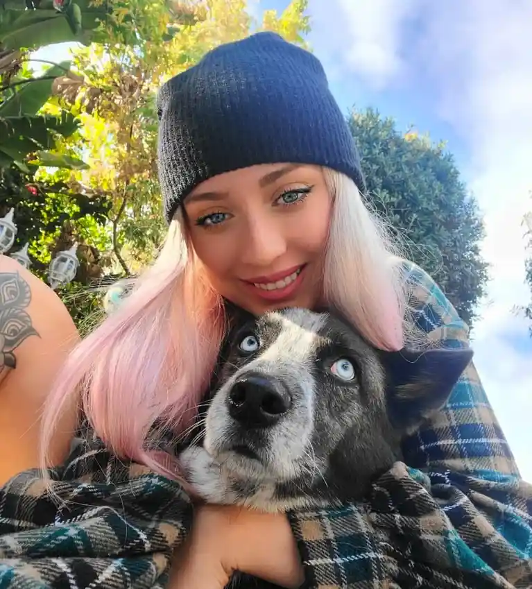 A young woman with pink hair and a dark beanie smiles while holding a black and white dog with striking blue eyes. The dog…