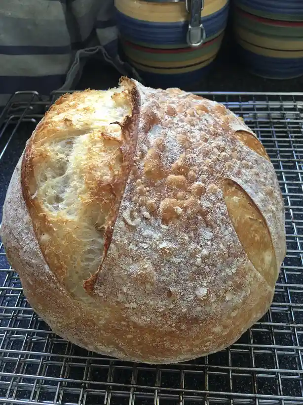 A round loaf of artisan bread sits on a metal cooling rack, its crust split open to reveal a golden, airy interior. The…