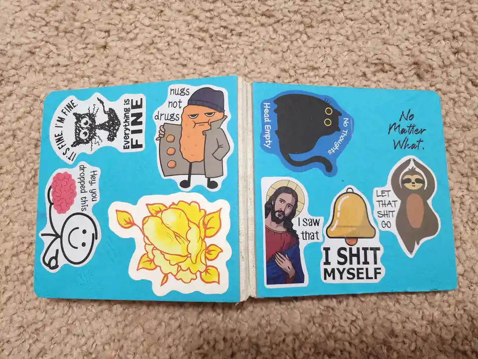 An open blue notebook or journal lies on a beige carpet, covered with various cartoon stickers. The stickers include a…