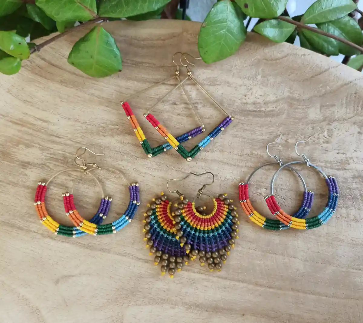 Four pairs of colorful, rainbow-patterned beaded earrings are displayed on a light wooden surface, partially framed by green…