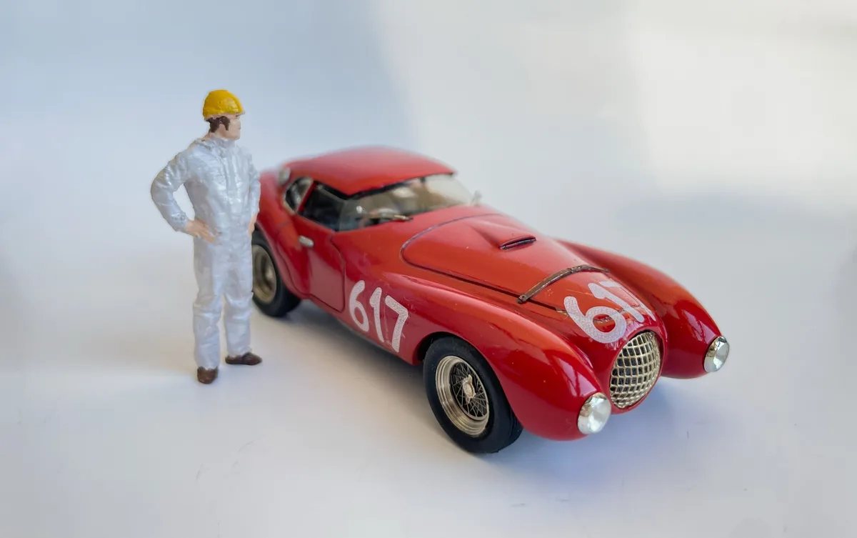 A small figurine of a man in a white jumpsuit and yellow hard hat stands confidently next to a red vintage race car model…