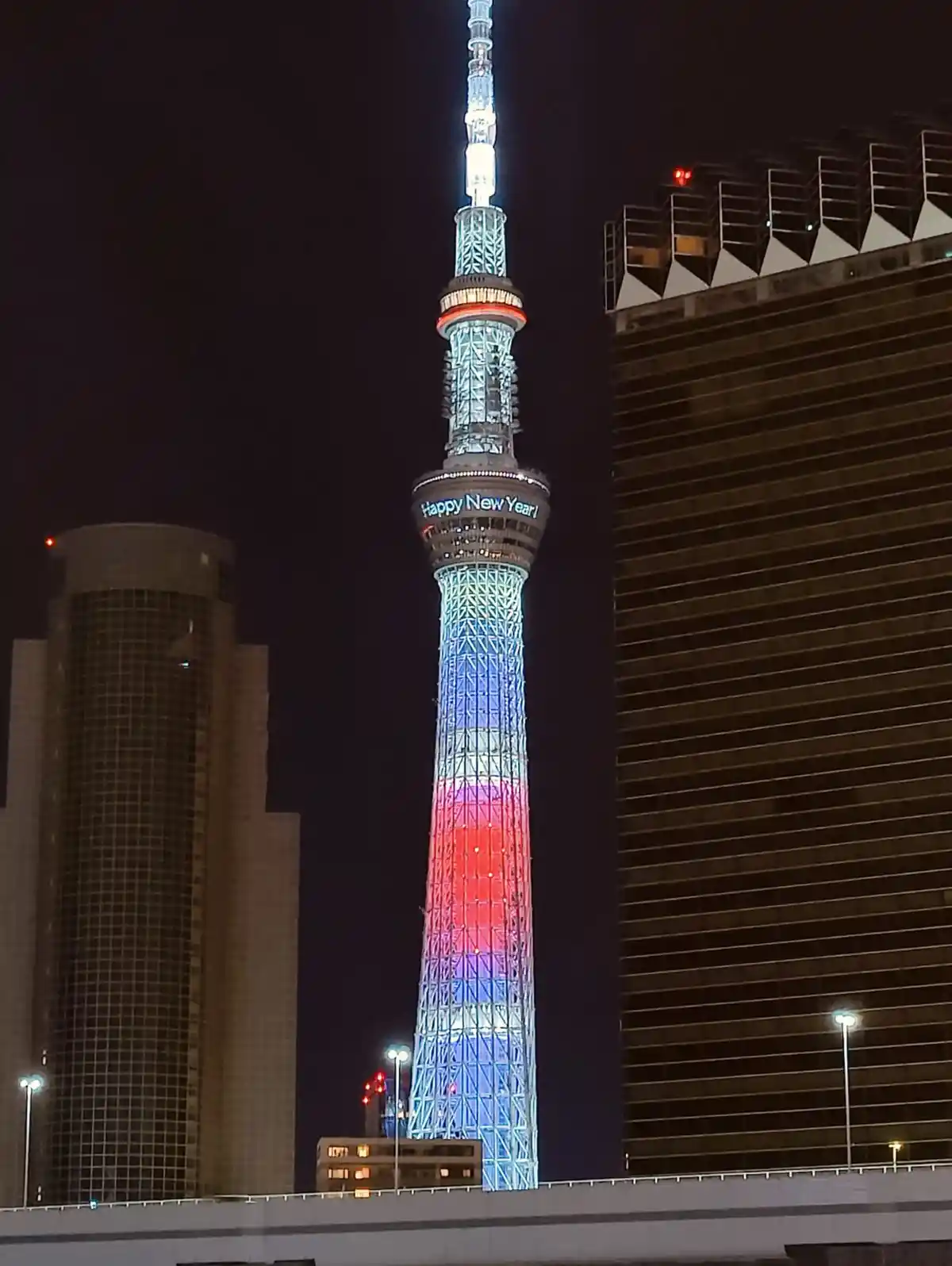 A nighttime photo of the Tokyo Skytree illuminated with red, white, and blue lights, with "Happy New Year!" displayed on its…