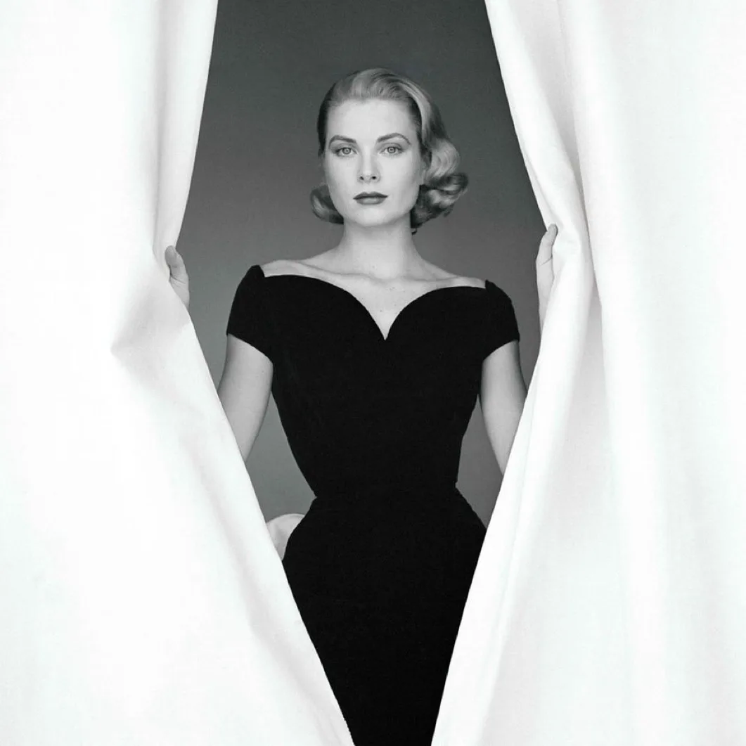 a black and white photograph shows actress grace kelly standing behind parted white curtains, wearing a black…