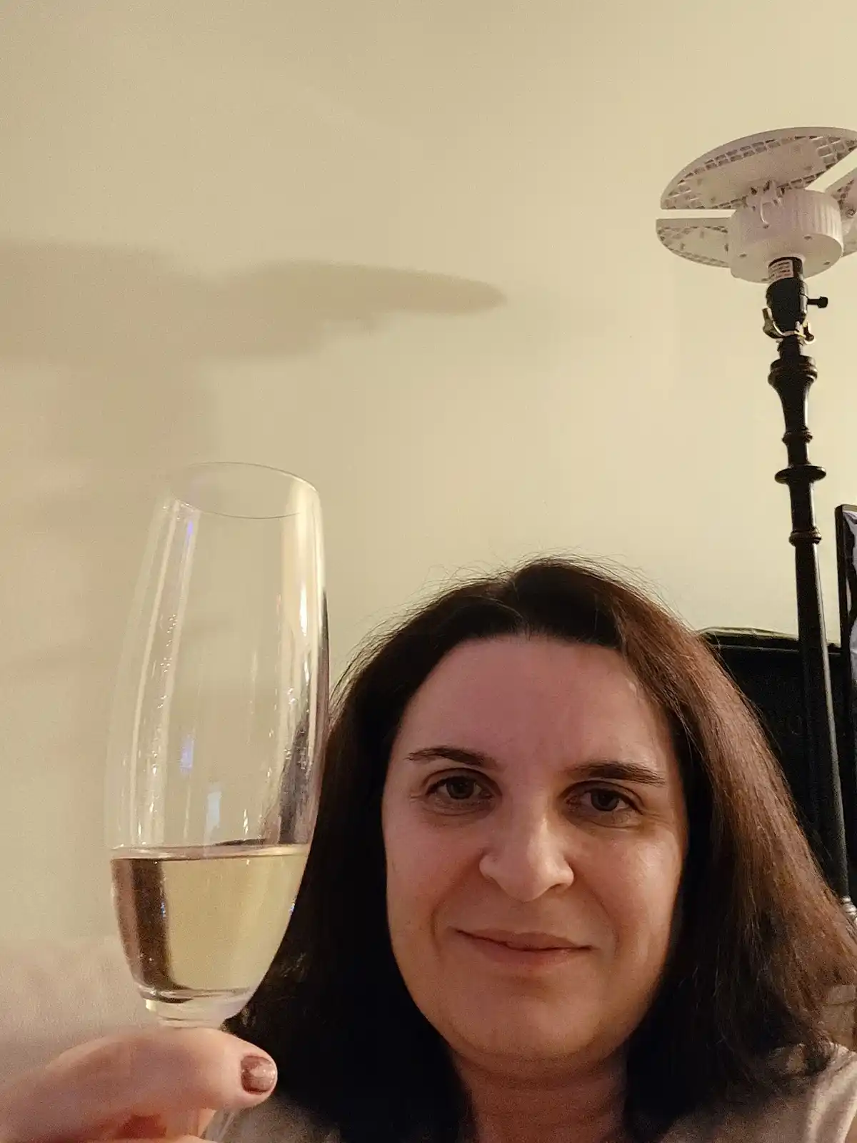 A woman with dark hair holds up a champagne flute containing a small amount of pale yellow liquid, looking directly at the…
