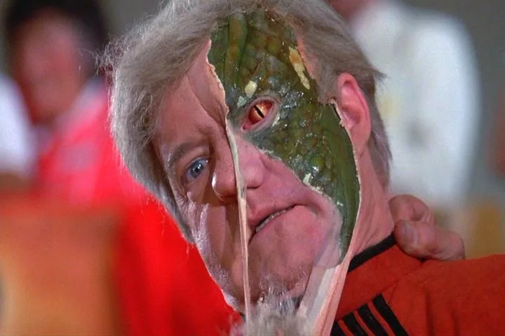 a man with gray hair and a red shirt has half his face covered in green reptilian skin with a glowing red eye, while the…