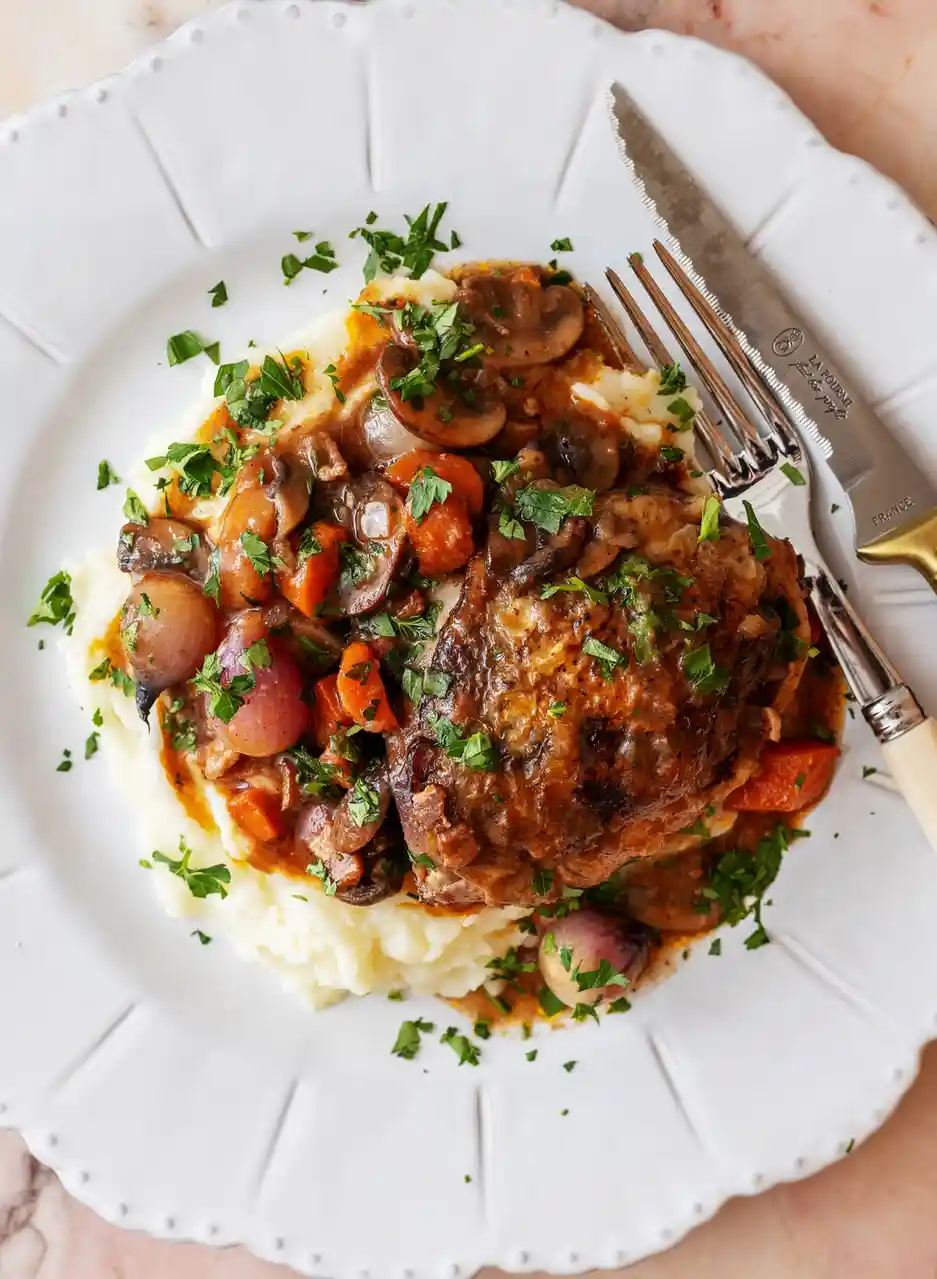 A top-down photo of a plated meal featuring a large piece of meat in a rich brown sauce with mushrooms, carrots, and pearl…