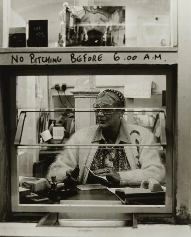 A black-and-white photo shows an older woman with glasses and a headband sitting behind a glass counter in what appears to…
