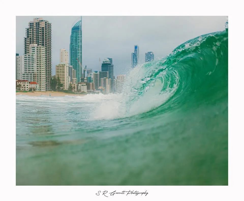 A low-angle photo of a large, curling green ocean wave in the foreground with a city skyline of tall buildings in the…