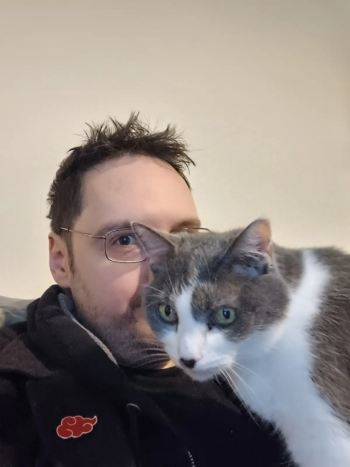 A man with glasses and messy hair takes a selfie while holding a gray and white cat that is resting its head on his…