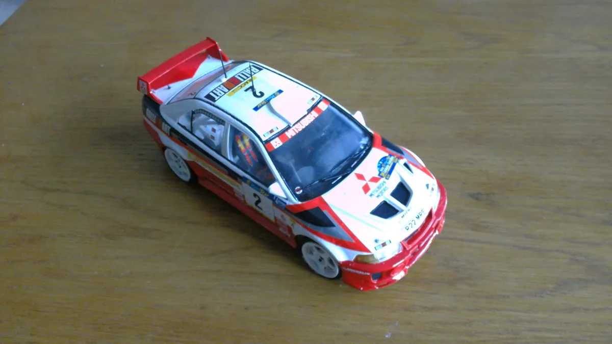 A red, white, and black die-cast model of a Mitsubishi rally car, number 2, sits on a wooden surface. The car features…