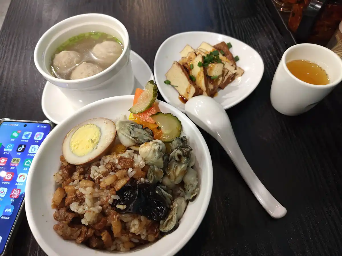 A table setting features a bowl of rice topped with oysters, a halved boiled egg, pickled vegetables, and braised pork…