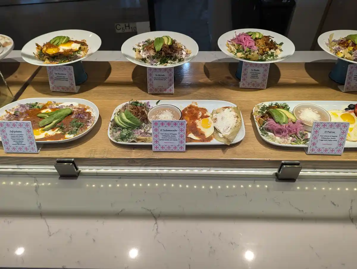 A display case in a restaurant shows multiple plates of colorful Mexican breakfast dishes like chilaquiles, huevos…