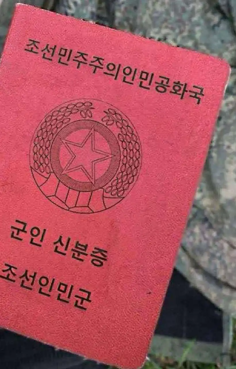 A close-up photo shows a red Korean People's Army ID card held in front of a person wearing camouflage clothing. The card…