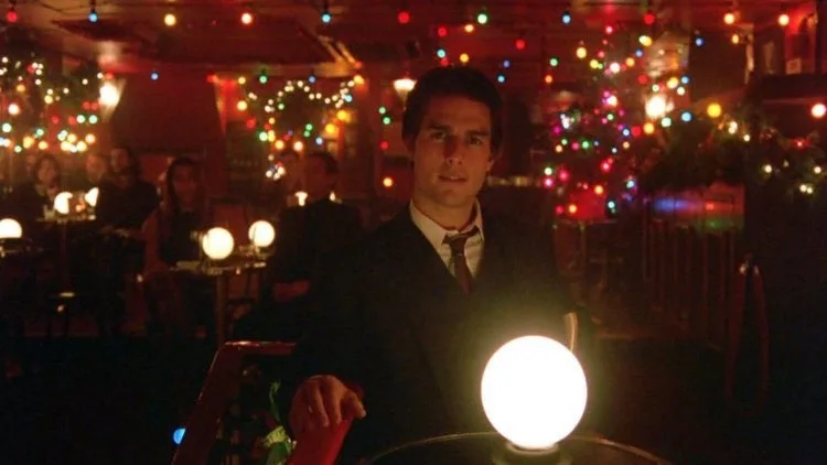 a man in a suit sits at a dimly lit table in a festive, bokeh-lit restaurant, looking directly at the camera with a serious…