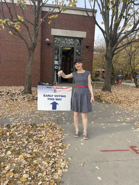 A woman in a polka-dot dress stands outside a brick building holding a sign that reads 'Mercer County EARLY VOTING THIS WAY'…