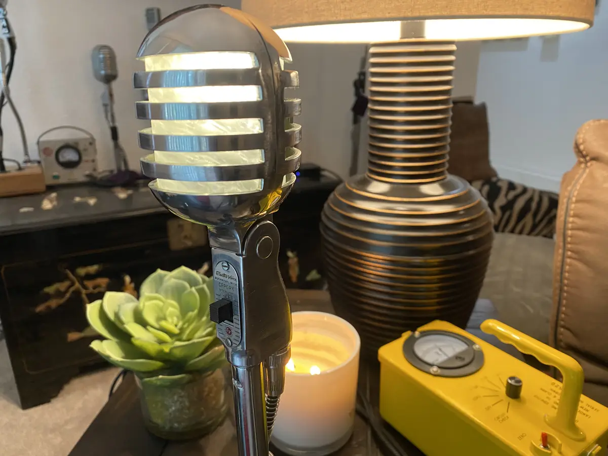 A vintage-style microphone with a glowing light inside its grille is placed on a table next to a lit candle, a potted…