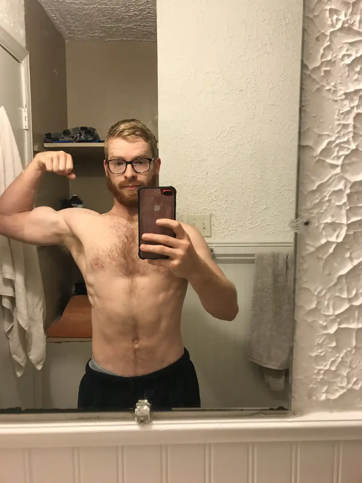 A shirtless man with glasses and a beard takes a mirror selfie in a bathroom, flexing his bicep while holding a smartphone…