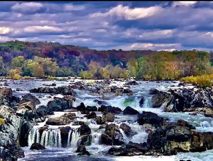 A scenic photo of a turbulent river flowing over dark rocks under a cloudy sky, with trees on the banks. The overlaid text…