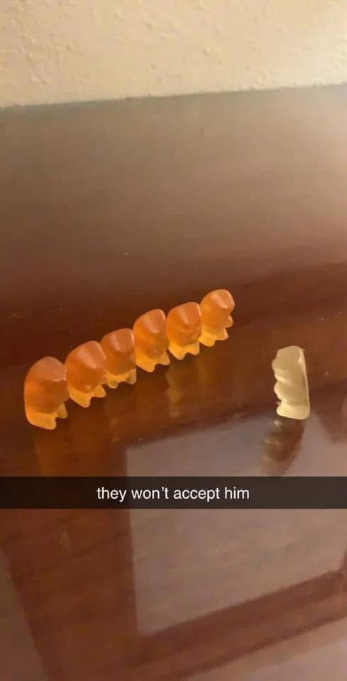 Five orange gummy bears stand in a line on a wooden surface, facing a single pale yellow gummy bear standing alone nearby. A…