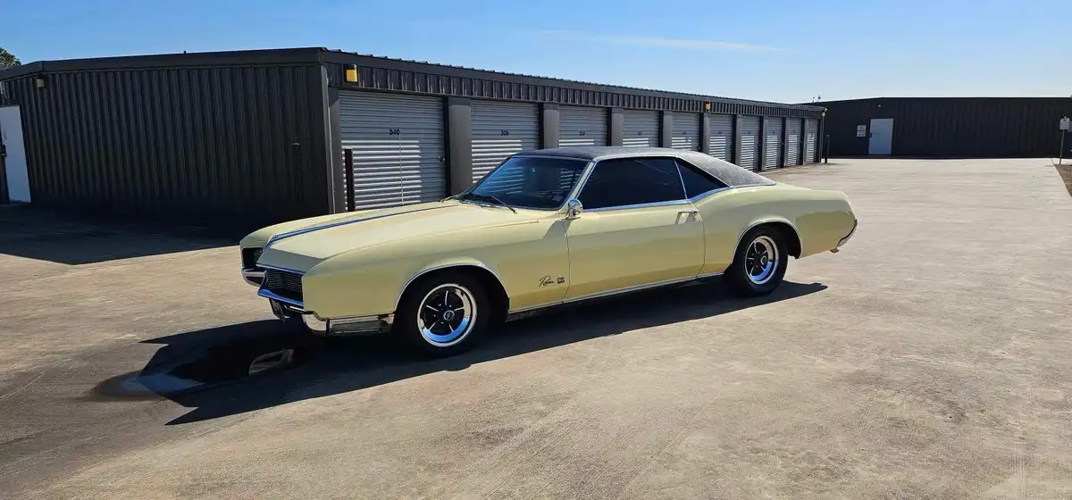 A pale yellow classic 1960s Buick Riviera GS muscle car is parked in front of a row of dark gray metal storage units with…