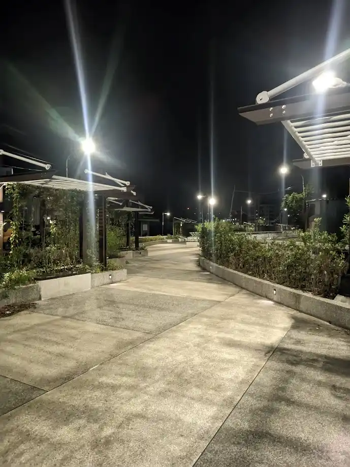 A nighttime photo of a well-lit, empty concrete walkway flanked by greenery and modern structures, with bright streetlights…