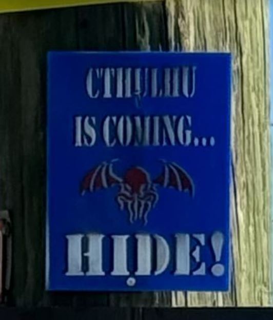 A blue sign with raised white lettering reads 'CTHULHU IS COMING... HIDE!' and features a red and white stylized bat-like…