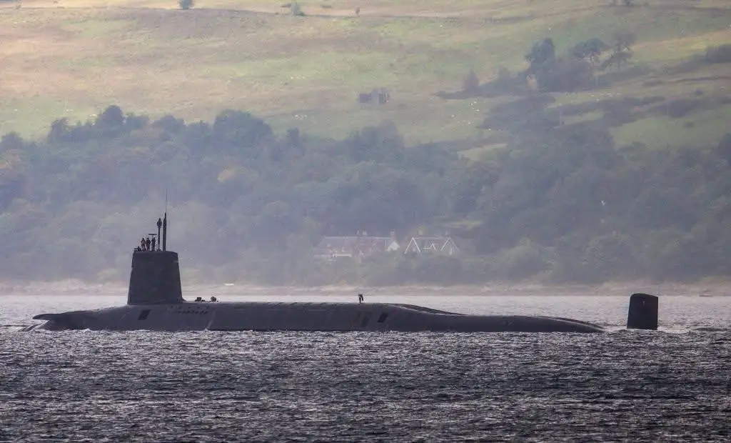 A dark grey submarine is partially surfaced in calm water, with its conning tower and periscope visible. Several people…