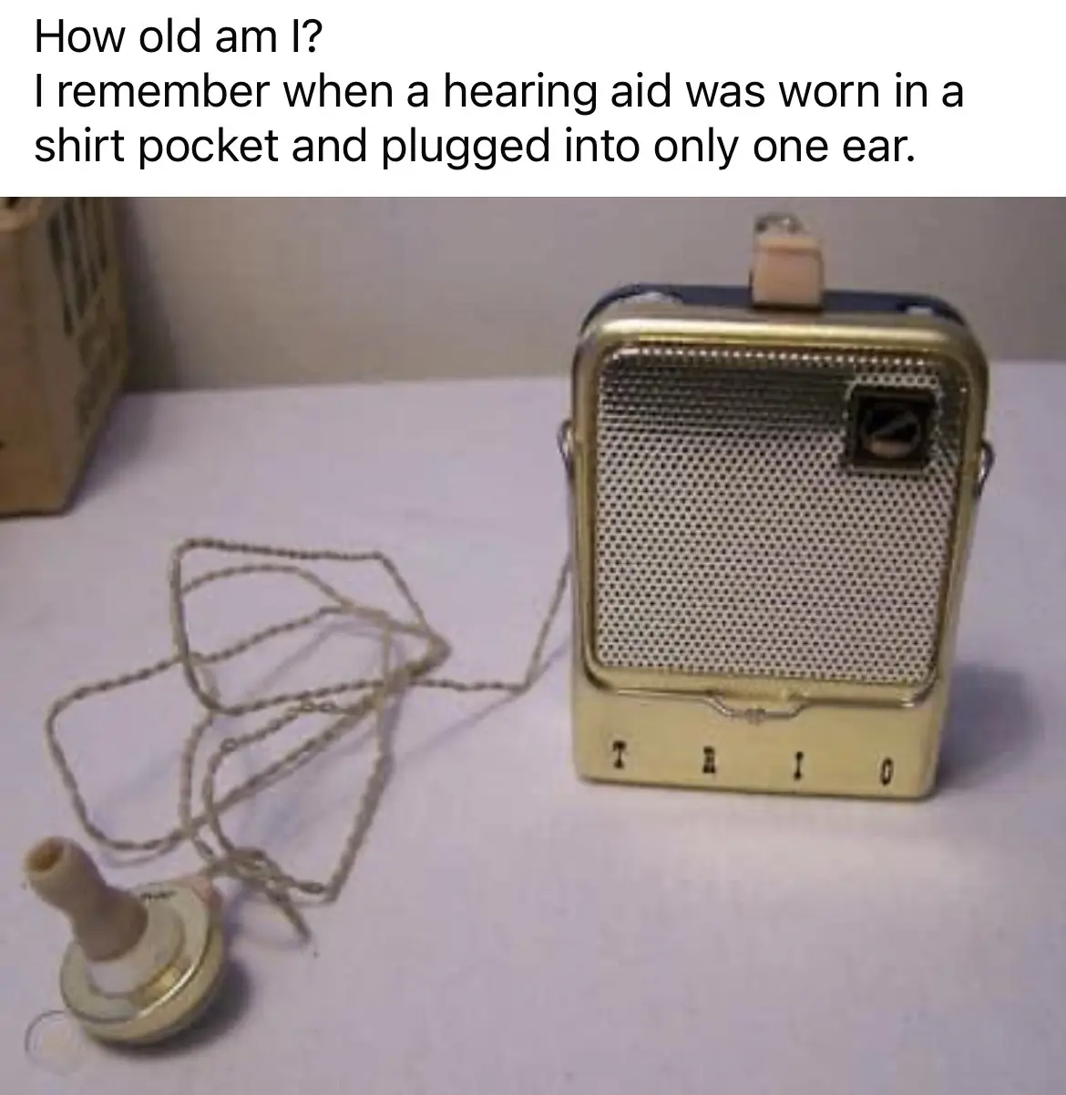 A vintage, beige, rectangular 'TRIO' brand hearing aid with a mesh speaker grille and a coiled wire connecting to a small…