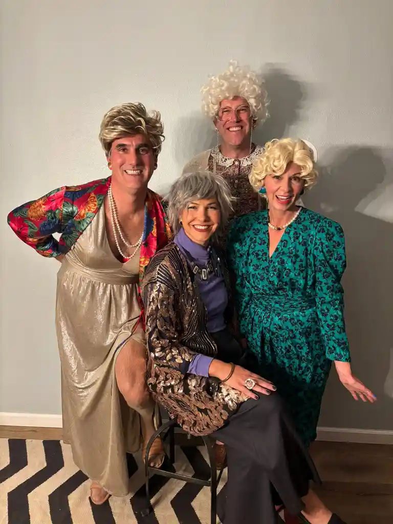four people dressed in exaggerated 1980s fashion, including big blonde wigs, sequined outfits, and pearl necklaces, posing…