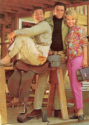 Three people pose together in a workshop setting. The man on the left is sitting on a saddle mounted on a wooden stand…