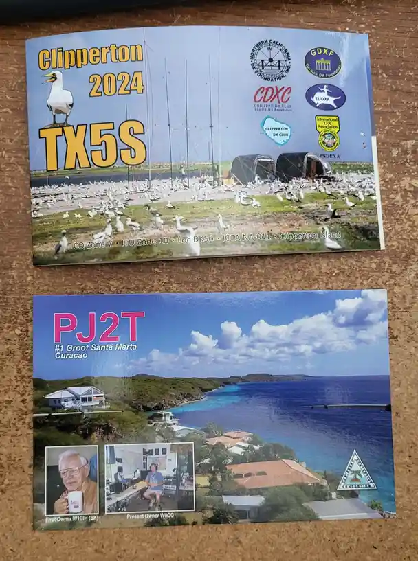 Two amateur radio QSL cards are displayed side-by-side on a corkboard. The top card features a photo of a remote island…
