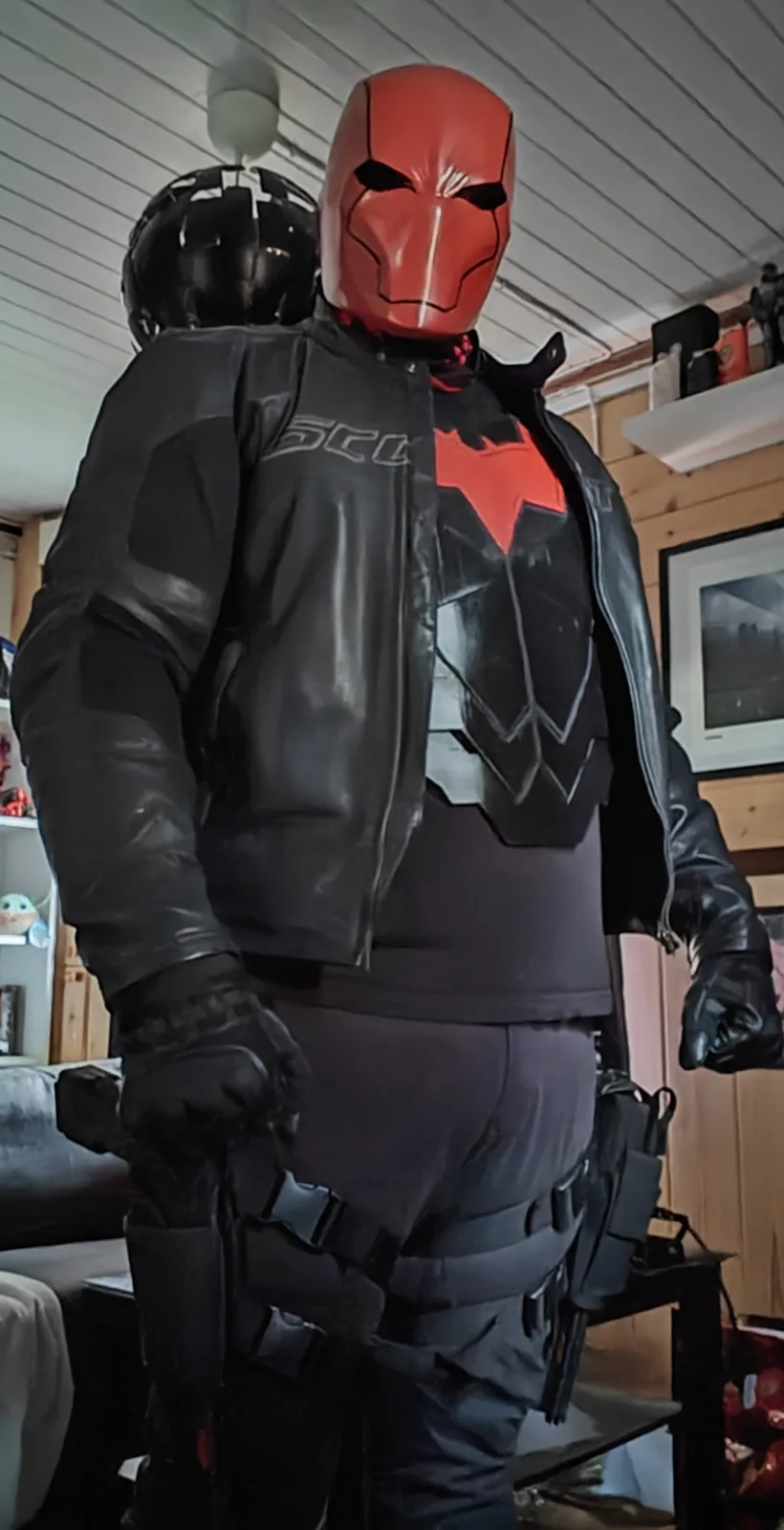 A person wearing a red, full-face mask and a black tactical outfit with a red bat symbol on the chest stands indoors. They…