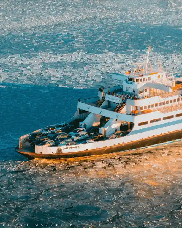 A white ferry named 'CAPE HENLOPEN' navigates through icy waters, its car deck loaded with vehicles, surrounded by…