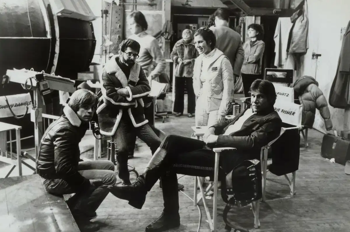 A black-and-white behind-the-scenes photo from the Star Wars set shows director George Lucas standing with arms crossed…