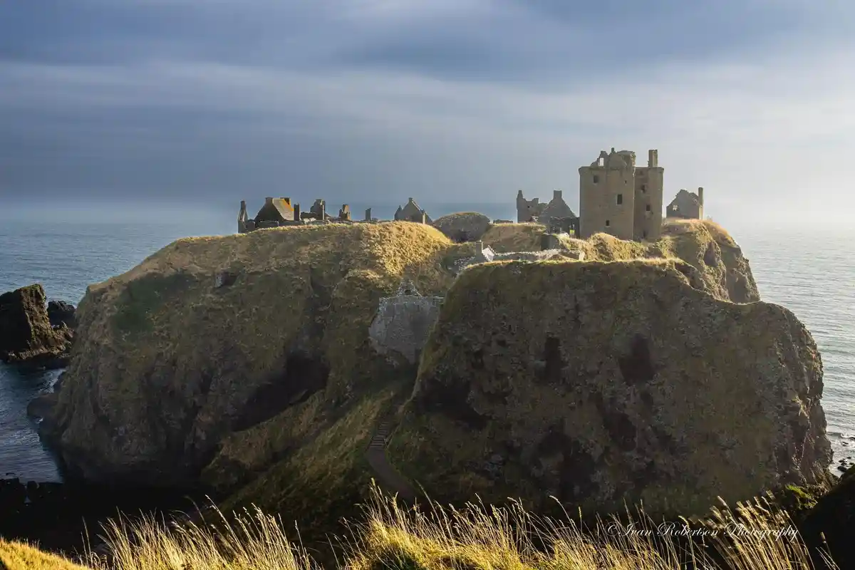 A scenic photograph shows the ruins of a stone castle perched atop a grassy, rocky cliff overlooking the ocean under a…