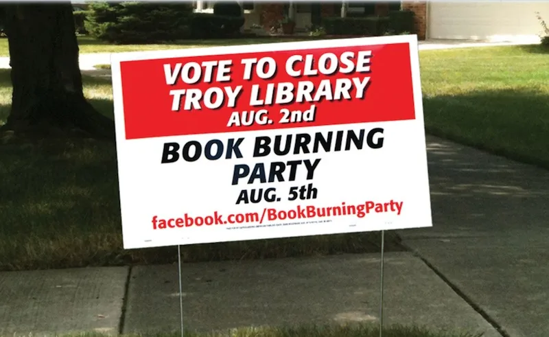 A red and white yard sign stands on a sidewalk in front of a house, advertising a 'VOTE TO CLOSE TROY LIBRARY' on August…