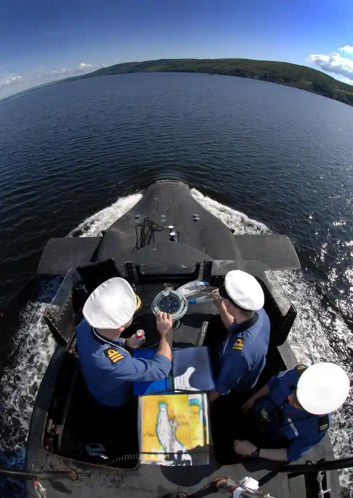 Three Royal Navy sailors in blue uniforms and white hats are on the bow of a black submarine, looking at a compass and a…