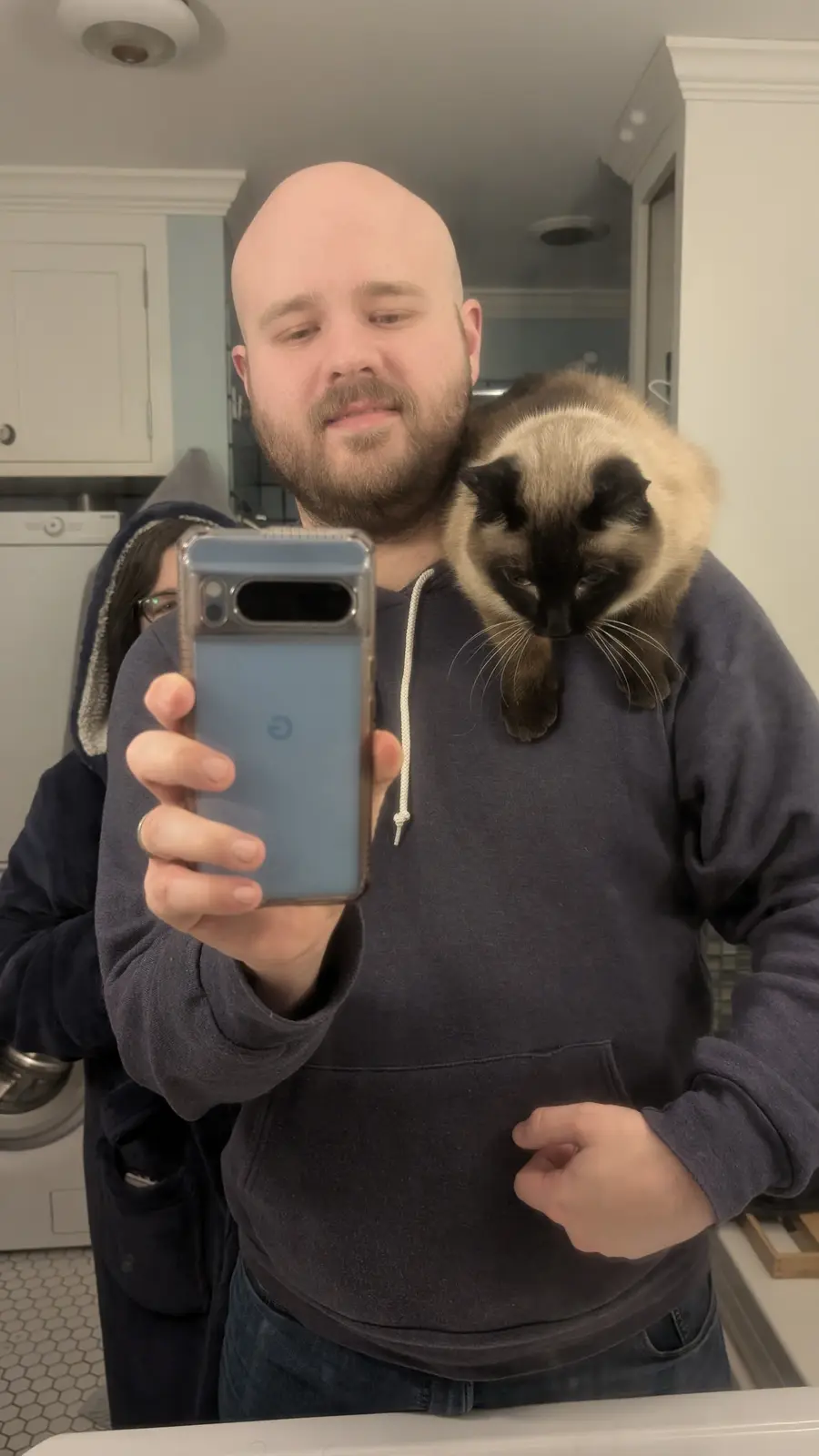 A bald man with a beard in a dark hoodie takes a mirror selfie holding a smartphone, with a siamese cat perched on his…