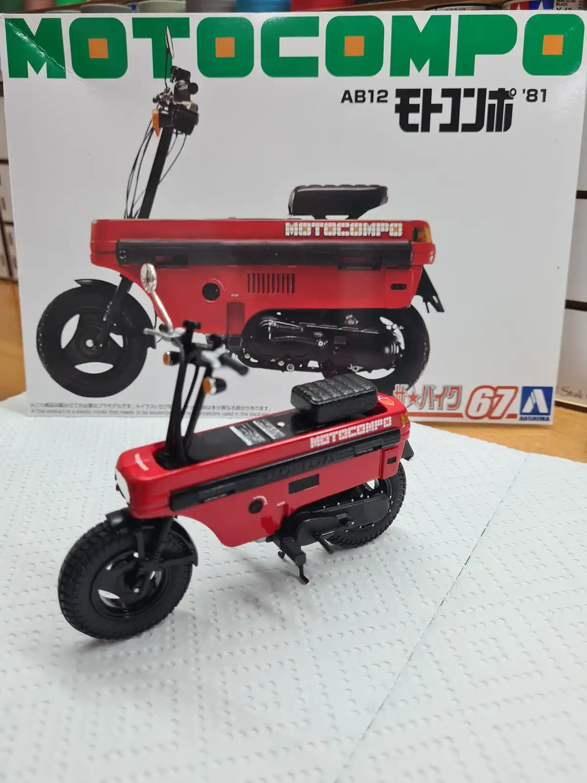 A red, assembled plastic model of a Honda Motocompo scooter sits on a white textured surface in front of its original box…