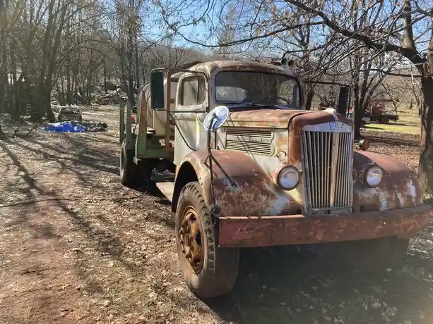 A heavily rusted, vintage white and red truck with a large grille and round headlights is parked on a dirt path surrounded…