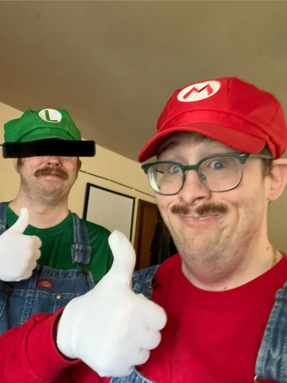 Two men dressed as Mario and Luigi from Super Mario Bros. pose for a selfie, both giving thumbs up. The man in the red Mario…