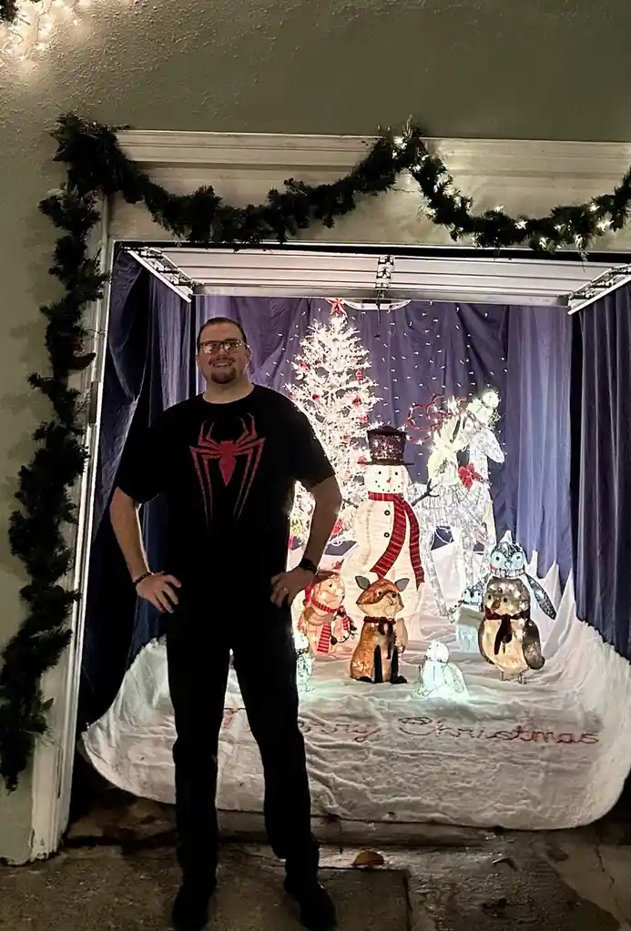 A man in a black t-shirt with a red spider logo stands confidently in front of a Christmas photo booth backdrop. The…