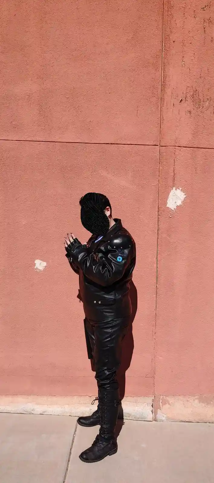 A person in full black leather outfit with elbow pads and tall boots stands against a pinkish wall, hands clasped in prayer…