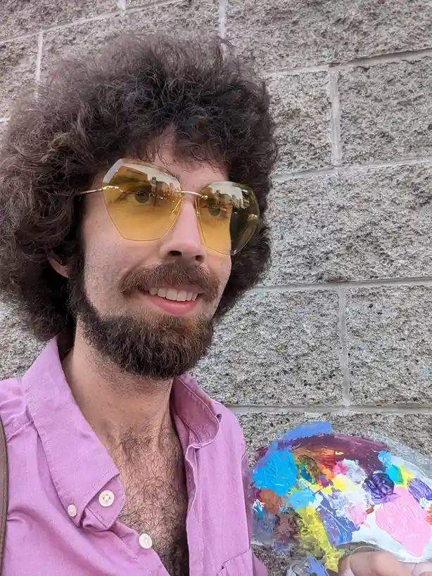 A man with curly brown hair, a beard, and large yellow-tinted sunglasses smiles while holding a paint palette filled with…