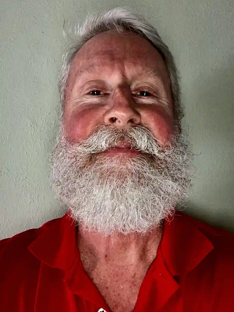 A close-up photo shows an older man with a full, white beard and mustache, wearing a red collared shirt against a plain…