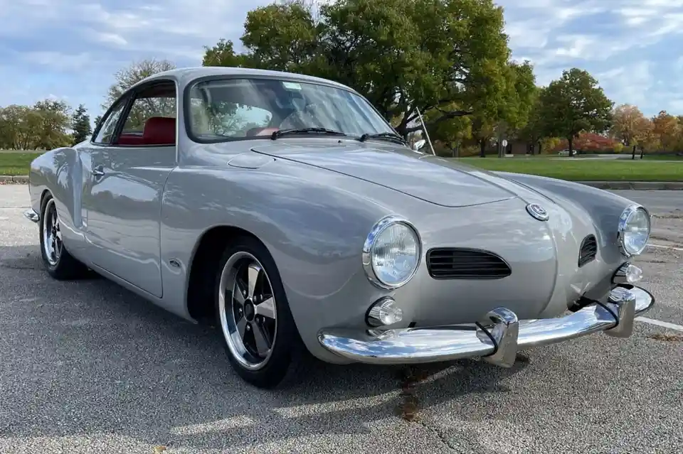 A silver vintage Karmann Ghia sports car is parked outdoors on asphalt with trees and a partly cloudy sky in the background…