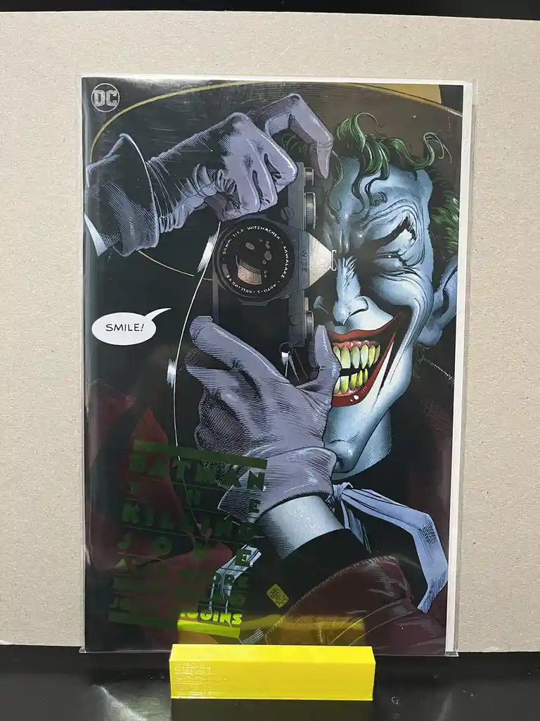 A comic book cover of 'Batman: The Killing Joke' shows the Joker holding a camera to his face, smiling maniacally, with a…